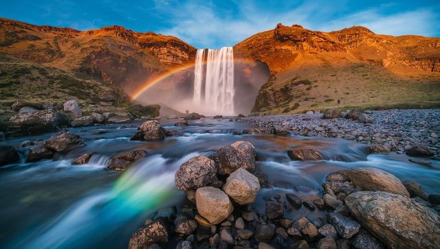 Golden-hour waterfall cascading into river valley with rainbow arc and rocky foreground
