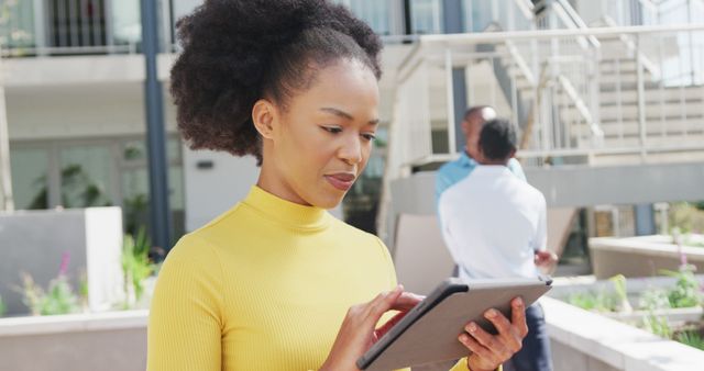 Professional woman with afro hair using tablet outside an office building. Ideal for business, technology, and modern work themes. Can be used for promoting digital tools, remote work, and professional settings.