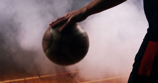 This image features a basketball player handling the ball on a smoky indoor court, with a focus on the hand and ball. Perfect for sports advertisements, motivational posters, and promotions related to basketball events or athletic products.