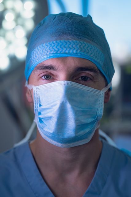 Male surgeon wearing surgical mask and blue scrubs in a hospital operating room. He is looking directly at the camera. This image is ideal for use in healthcare-related articles, medical blogs, hospital websites, and educational materials about surgery and healthcare professions.