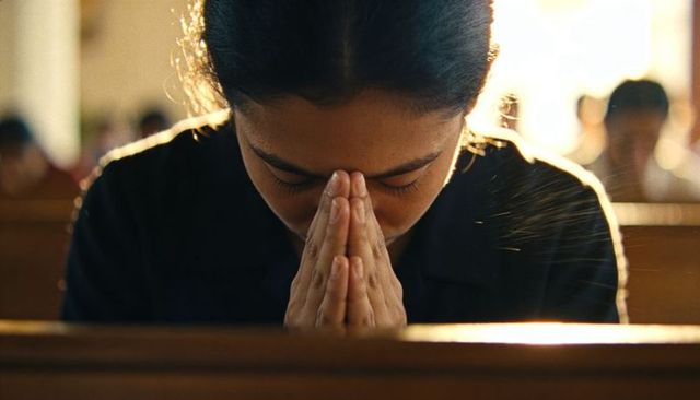 Praying asian woman sitting in church pew backlit by sunlight showing reverence and contemplation