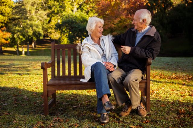 Elderly couple sitting on a wooden bench in a park, smiling and enjoying each other's company. The autumn foliage adds a warm, colorful background. Ideal for use in advertisements, brochures, or articles related to senior living, retirement, companionship, and outdoor activities.