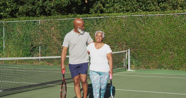 Senior couple smiling and holding tennis rackets while walking on tennis court. Ideal for promoting active lifestyles among older adults, health and fitness campaigns, retirement community advertisements, and all-age leisure activities. Emphasizes joy, companionship, and staying fit in the golden years.
