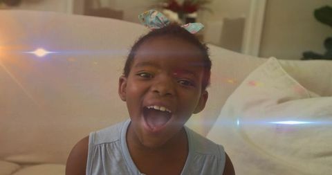 Laughing young girl with patterned bow sitting on beige sofa with playful lens flare