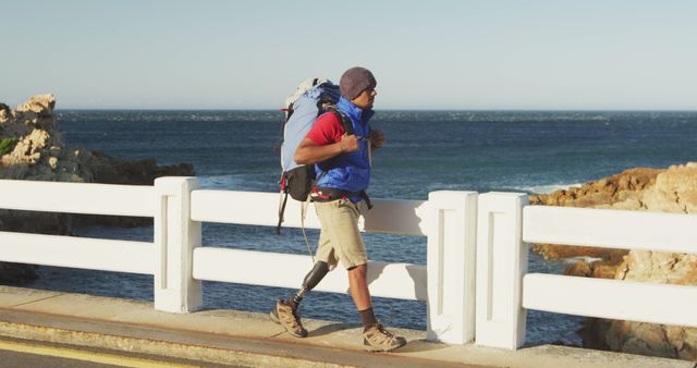 Biracial man with prosthetic leg trekking with backpack crossing bridge by the sea. Long distance walking, fitness, challenge, disability and healthy outdoor lifestyle.