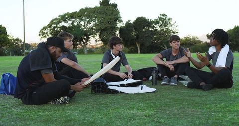 Cricket teammates discussing strategy after practice, five players sitting with bat and pads