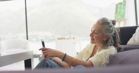 Mature woman relaxing on floor leaning against sofa using smartphone in sunlit living room