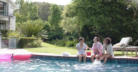 Diverse women sitting on pool edge splashing water, enjoying summer backyard party