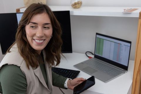 Woman working from home smiling while checking smartphone and laptop spreadsheet