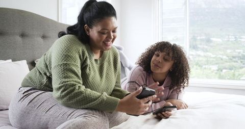 Friends sharing smartphones while reclining on sunlit bedroom bed, cozy modern lifestyle