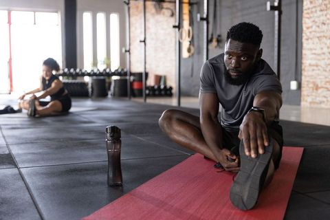 Diverse athletes stretching on mat in industrial gym with water bottle and smartwatch