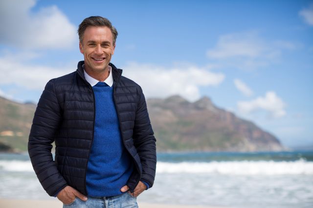 Mature man standing on beach, smiling confidently with ocean and mountains in background. Ideal for use in travel brochures, lifestyle blogs, vacation advertisements, and wellness articles.
