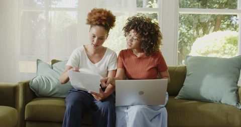 Two African American women reviewing documents with laptop on sofa for home collaboration