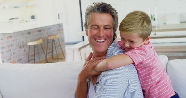 A Caucasian middle-aged man shares a joyful embrace with a young boy in a bright, modern living room, with copy space. Their laughter and close hug convey a warm, loving family moment.