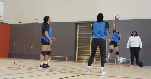 Diverse female volleyball team practicing spikes and drills in gym with coach supervising