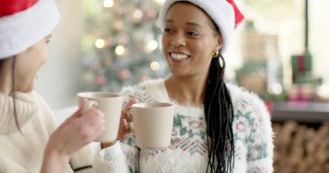 Diverse friends clinking ribbed mugs in santa hats and cozy sweaters by christmas tree