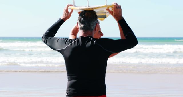 A middle-aged Caucasian man in a wetsuit carries a surfboard on his head at the beach, with copy space. His posture and attire suggest he is preparing for a surfing session in the ocean.