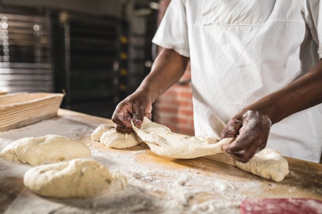 Midsection of african amerian male baker making dough at counter in bakery kitchen. unaltered, blue-collar worker, skilled, food and drink industry concept.
