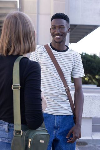 Young friends talking and smiling on urban campus courtyard with shoulder bags in daytime