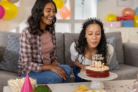 Teenage daughter blowing out 18th birthday candles while mother smiling on sofa
