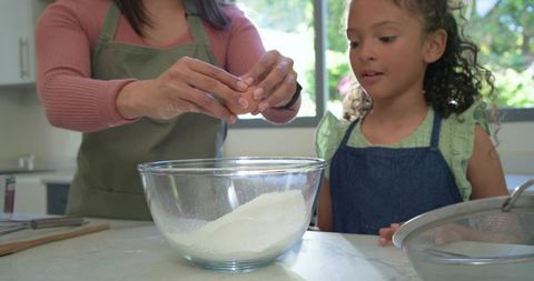 Mother and daughter cracking egg while preparing batter in bright home kitchen