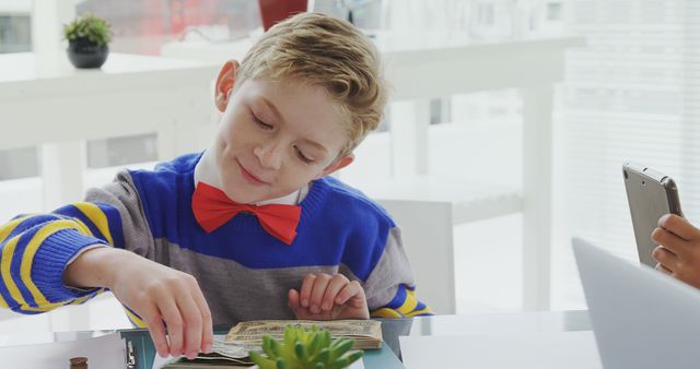 Child in colorful sweater and red bow tie counting money at office desk, a hand holding smartphone in background, represents future entrepreneurs, financial education. Useful for blogs about financial literacy, stock imagery for educational websites or articles indicating young entrepreneurship.