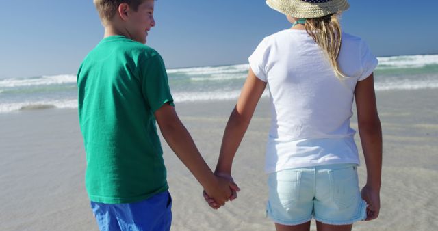 A young Caucasian boy and girl hold hands while walking on a sandy beach, with copy space. Their casual attire and the sunny beach setting suggest a relaxed and carefree day by the sea.