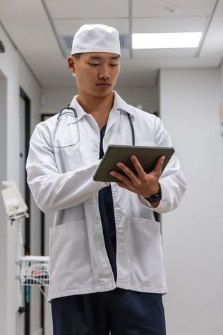 Asian male doctor standing in clinical corridor using tablet and wearing stethoscope