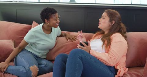 Multicultural friends chatting on pink sofa, laughing and holding smartphone