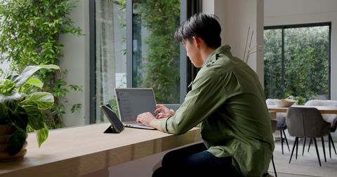 Asian Man Working on Laptop in Minimalist Home Office by Window