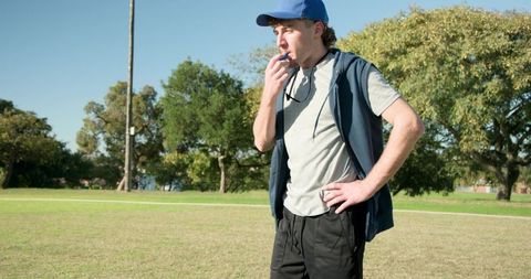 Coach blowing whistle during outdoor sports practice on grassy field wearing blue cap