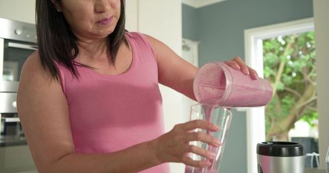Midlife asian woman pouring berry smoothie into glass in modern kitchen wearing pink tank