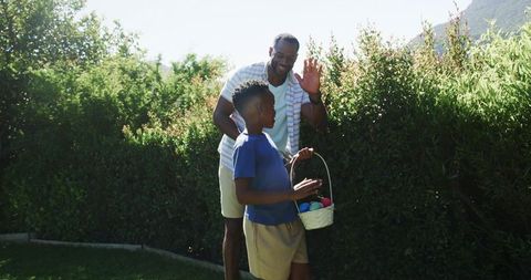 African american father waving, teenage son holding easter egg basket in sunlit backyard