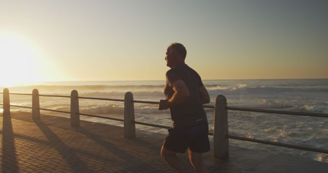 Mature man is running along the coast during a beautiful sunrise. The scene captures the man in silhouette against the backdrop of waves crashing on the shore. This can be used for promoting healthy lifestyle habits, fitness programs for seniors, or inspirational content about staying active and fit as one ages.