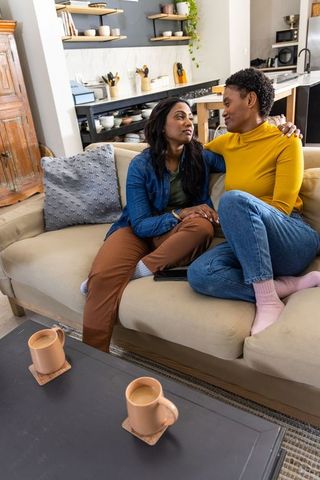 Affectionate black female couple cuddling on couch holding hands sharing tablet, cozy home