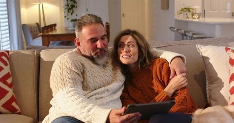 Middle-aged couple snuggling on beige sofa in cable-knit sweaters while holding tablet