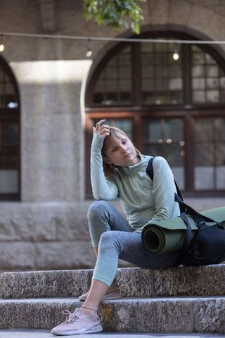Young woman sitting on stone steps holding yoga mat in urban campus courtyard, contemplative