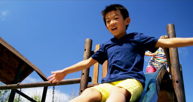 A young boy enjoying a sunny day at a playground, going down a blue slide with his arms spread wide. The background includes another child in colorful clothing and wooden playground structures. This image can be used for educational materials, summer camp promotions, childhood and parenting articles, and advertisements for outdoor play equipment.