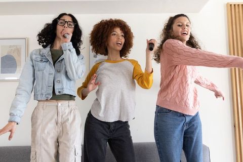 Diverse friends singing and dancing on sofa holding microphones in modern living room