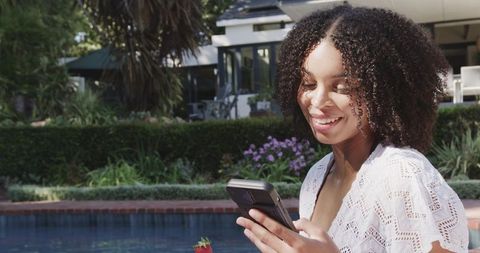 African american woman smiling while using phone poolside in lace coverup with strawberry