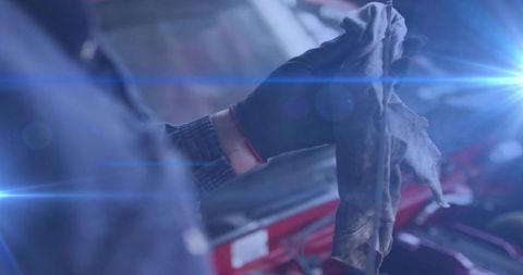 Female mechanic holding dirty rag and gloved hand near open red car hood and engine with lens flares
