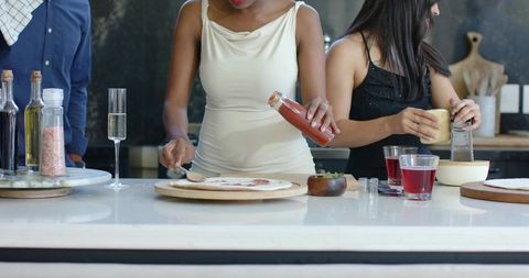 Friends preparing homemade pizzas in modern kitchen, pouring sauce and grating cheese