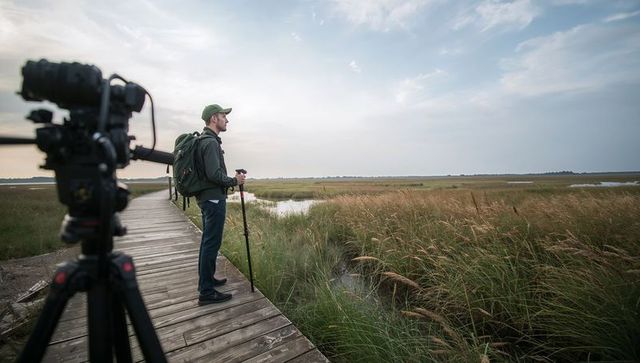 Photographer standing on boardwalk overlooking coastal marsh at sunrise, tripod ready