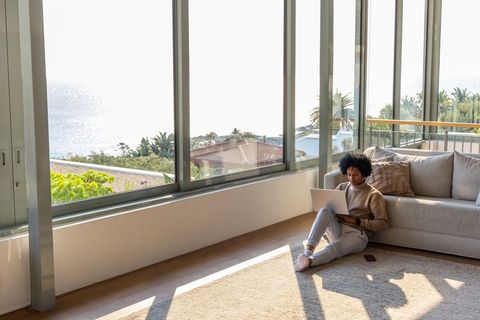African american woman working remotely on laptop in sunlit coastal living room with ocean view