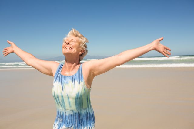 Senior woman standing on beach with arms outstretched, smiling joyfully under clear sky. Perfect for themes of happiness, freedom, active lifestyle, vacation, and relaxation. Ideal for travel brochures, wellness blogs, and retirement living advertisements.