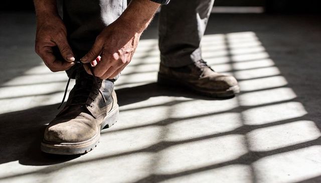 Worker tying worn leather work boots under dramatic slatted sunlight and strong shadows