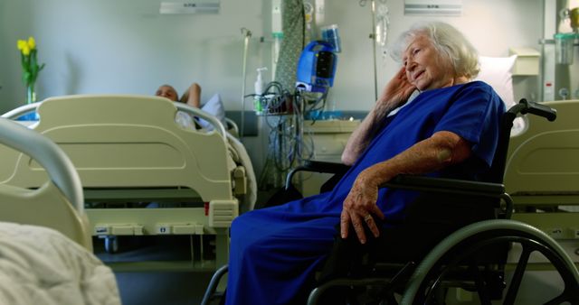 An elderly woman sitting in a wheelchair wearing a blue robe in a hospital room. The background shows hospital beds and medical equipment, indicating a healthcare setting. This can be used in articles, blogs, and websites discussing elderly care, health challenges, hospital care, patient experiences, or issues related to aging and medical services.