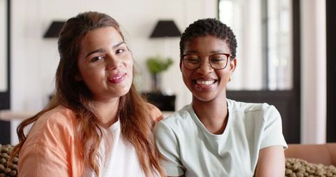 Smiling diverse friends sitting on cozy living room couch wearing tops, natural daylight