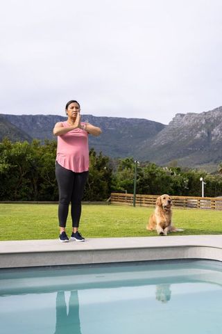 Mature woman practicing yoga prayer pose poolside with golden retriever and mountain view