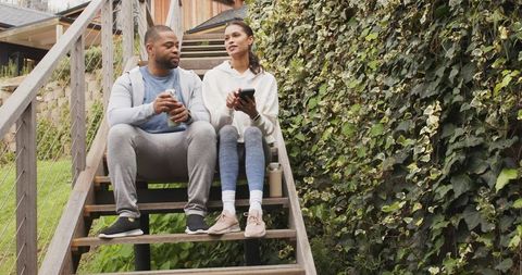 Couple sitting on wooden stairs talking over coffee while using tablet by ivy-covered deck